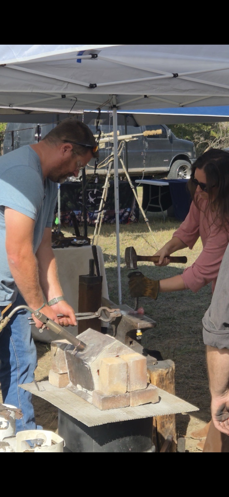 Two people working hot metal on an anvil under a tent