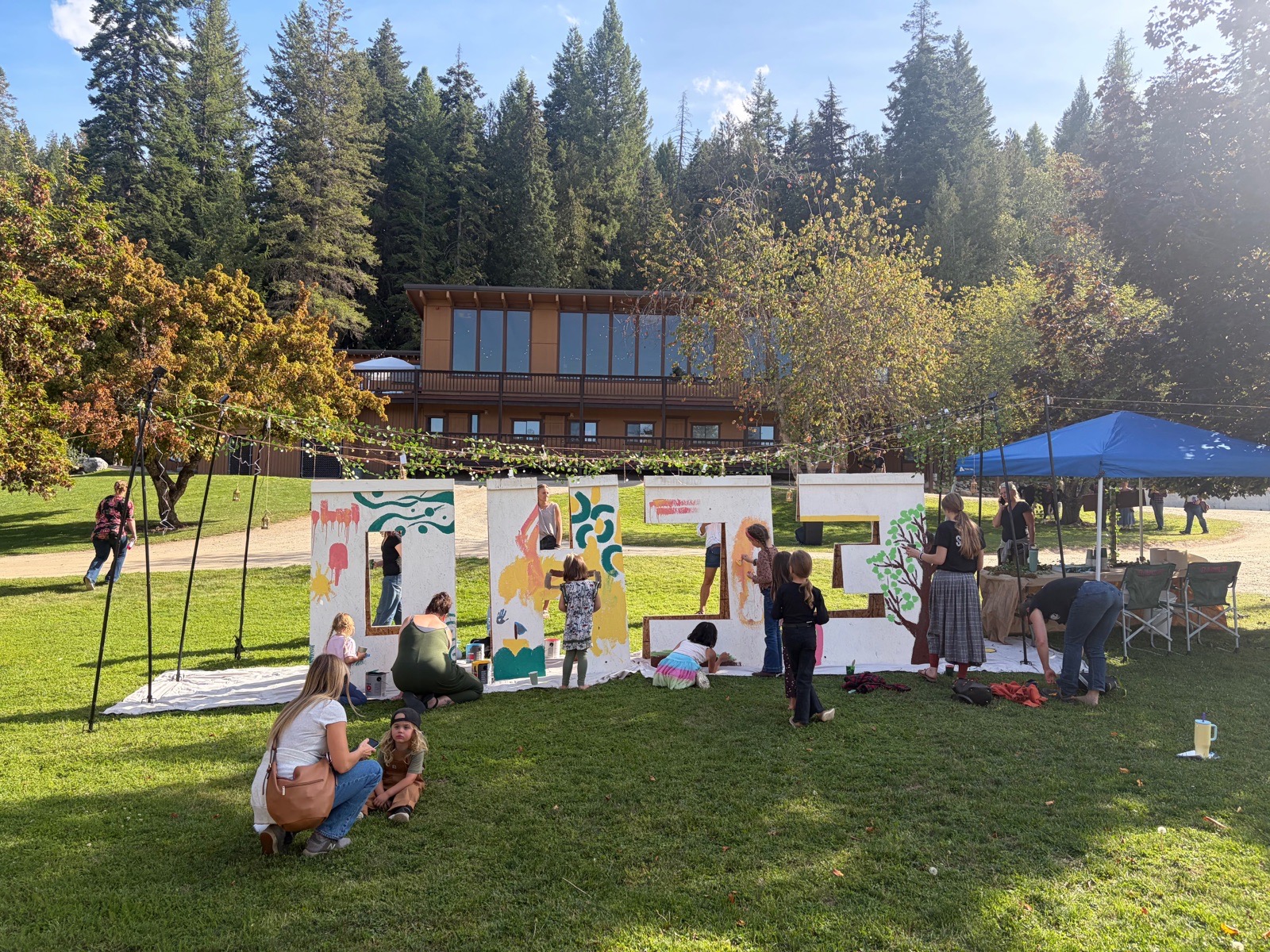 People painting tall panels on the lawn in front of the lodge, a tree turning orange at the left