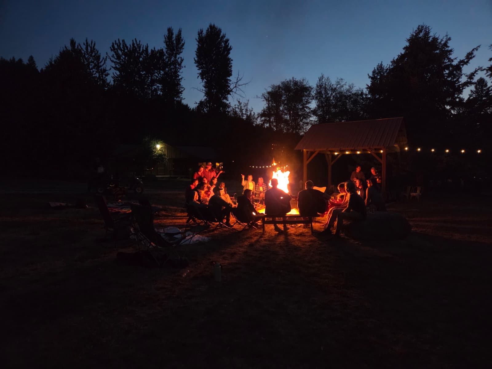 The fire circle at night, the crew gathered around it, string lights on the pavilion behind